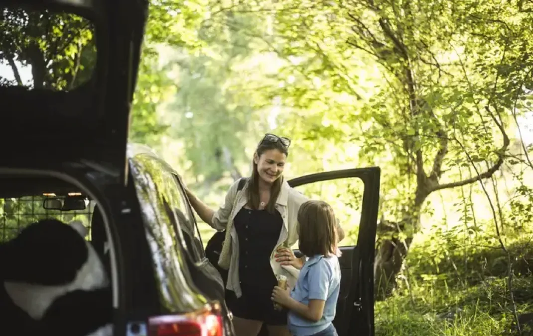 A mother is standing with her daughter between their Electric Vehicle and the open driverside door. They are looking at each other and the mother has one hand on the car. The car is black and the car boot is open and it parked in a forrest.