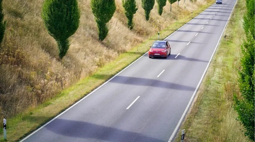 A red car driving on a road lined with trees, on a bright day.