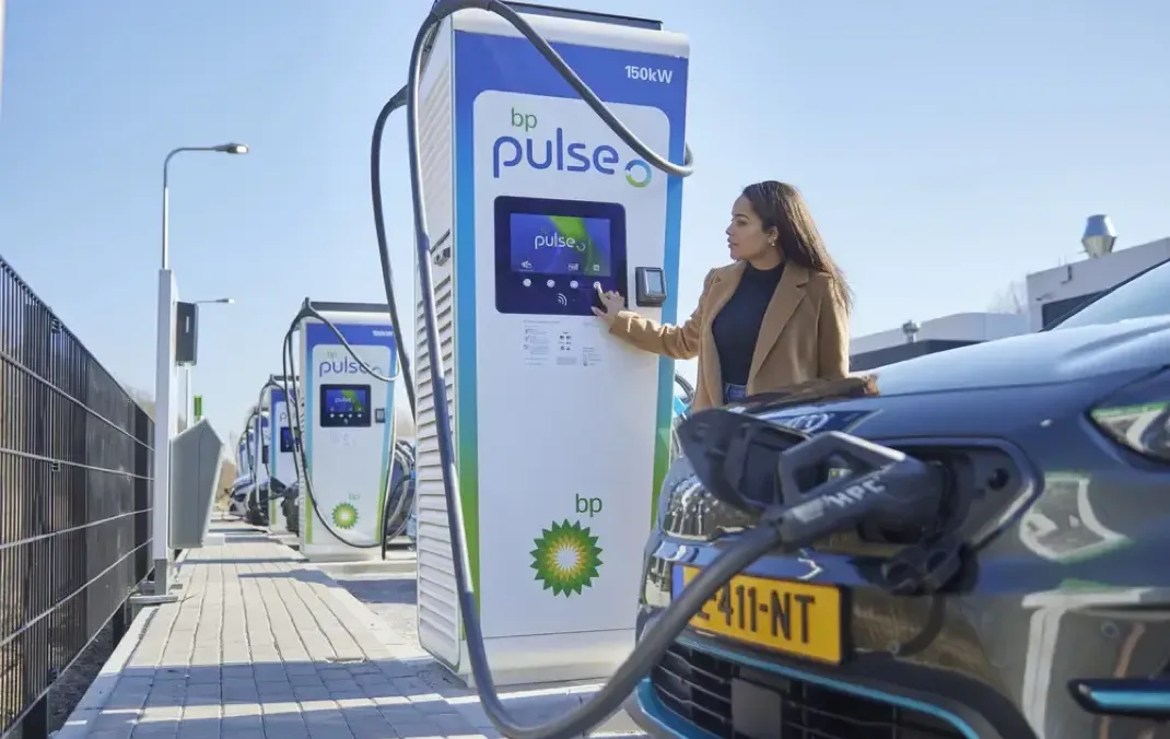 An electric vehicle parked in a charging bay, one of several in a row, connected to a 150kW charger. A woman is standing next to the vehicle with one hand on the charger's interface. The scene is set in a clean, modern charging area under a clear blue sky.
