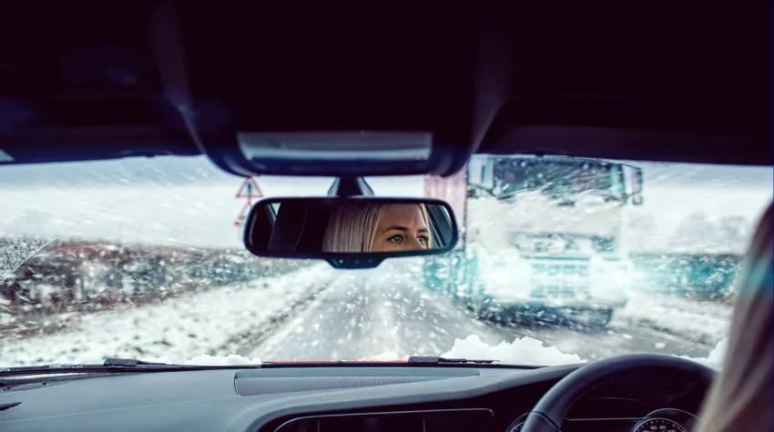 View from inside a car during a hailstorm, looking through the windscreen as hailstones strike the glass. An oncoming truck is visible on the road ahead, and the reflection of woman driving can be seen in the rearview mirror.