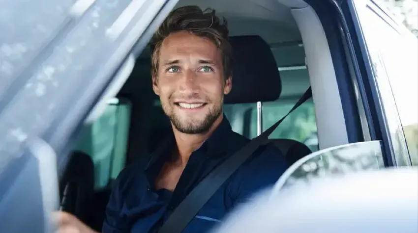 A man in a blue shirt is sitting in the driver seat of his Electirc Vehicle and looking out of the window smiling. The window is wound down.