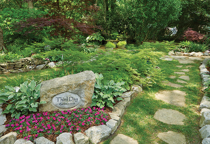 Garden stone ornament: Shade-tolerant annuals supply colorful accents in beds throughout the garden. Bright pink impatiens (*Impatiens* hybrd) in this spot provide a colorful skirt for the stone that bears the garden’s name and attracts attention to the path.
