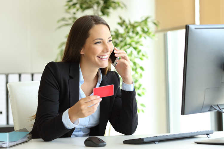Woman in office smiling on phone holding business card looking at computer screen