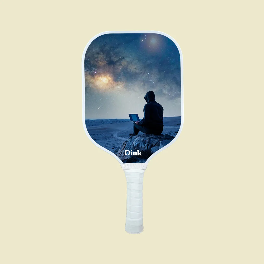A custom pickleball paddle from Dink with a man sitting under a starry night with the laptop