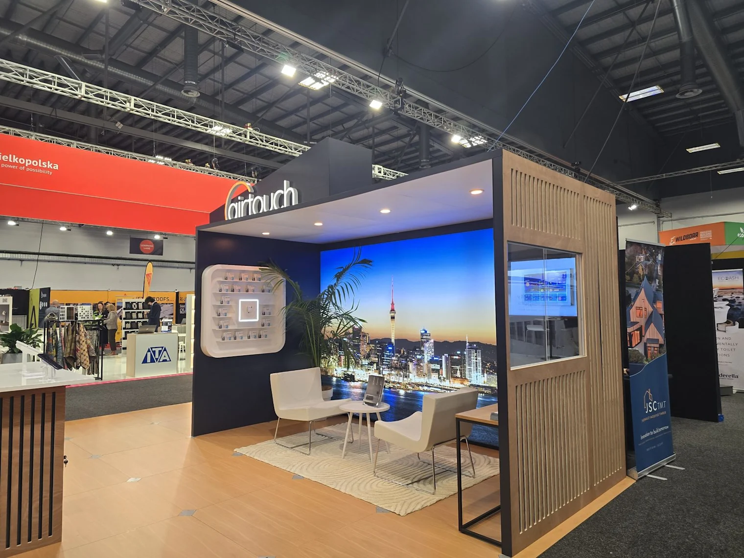 The lounge and seating area of the AirTouch stand at BuildNZ 2026, with the Auckland night-skyline screen.
