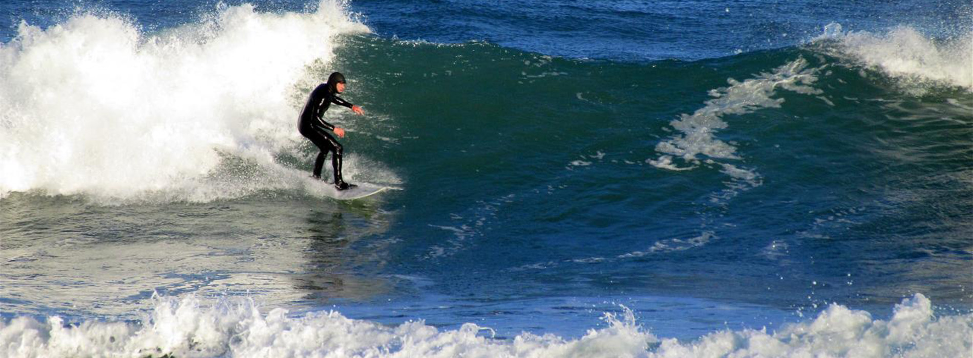 A surfer in a black wetsuit rides along the face of a large blue-green wave, with white foam crashing behind them.