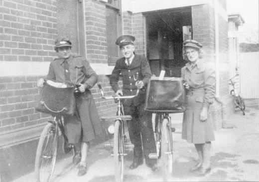 Three people wearing postal uniforms standing next to circa 1940s era bicycles.