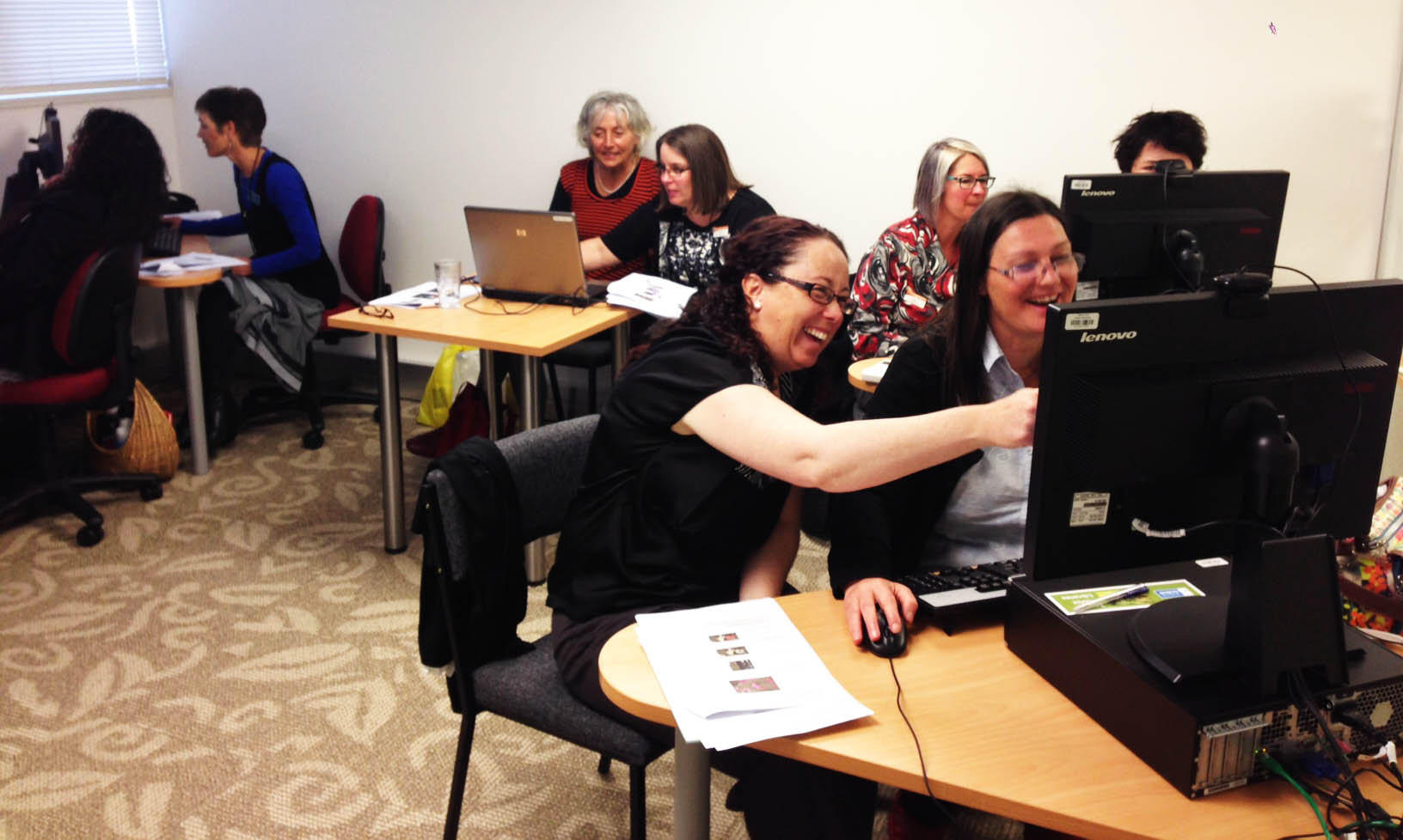 A group of people smiling as they work in pairs at computers.