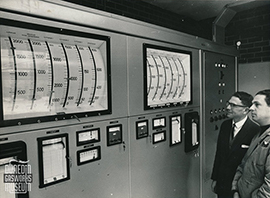 Two individuals standing in front of a large control panel with multiple gauges, meters, and two large curved scale displays.