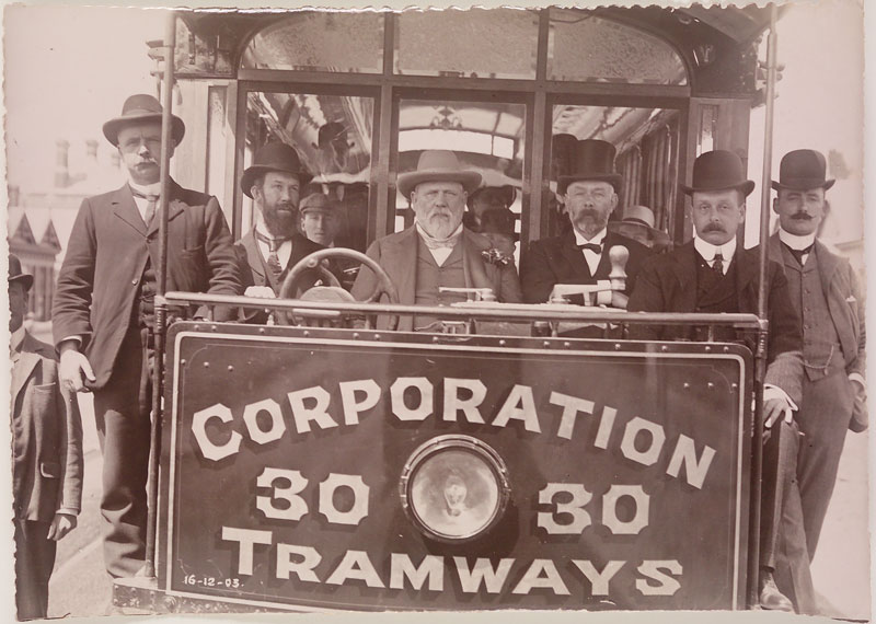 Six men wearing three piece suits and hats standing in the driver's area of a tram. The tram is painted with the text 'Corporation 30 30 Tramways'.