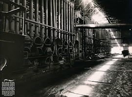 Interior view of an industrial retort house with rows of large cylindrical retorts and pipes along the wall, illuminated by overhead light streaming through the roof.