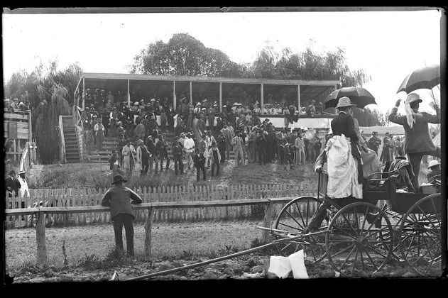 A crowded grandstand and people holding umbrellas standing on a horse and carriage.