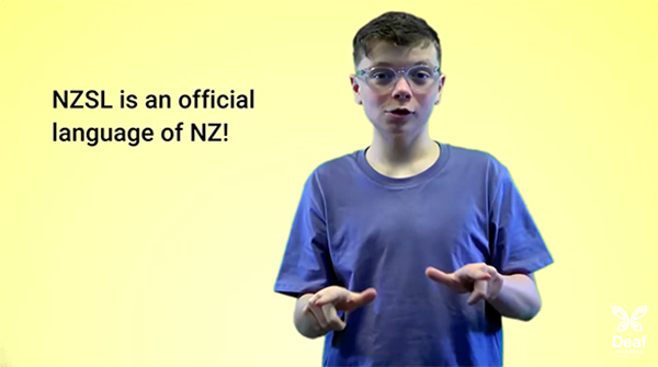 A person wearing a blue t-shirt is signing in New Zealand Sign Language against a bright yellow background. Text on the left reads: 'NZSL is an official language of NZ!'