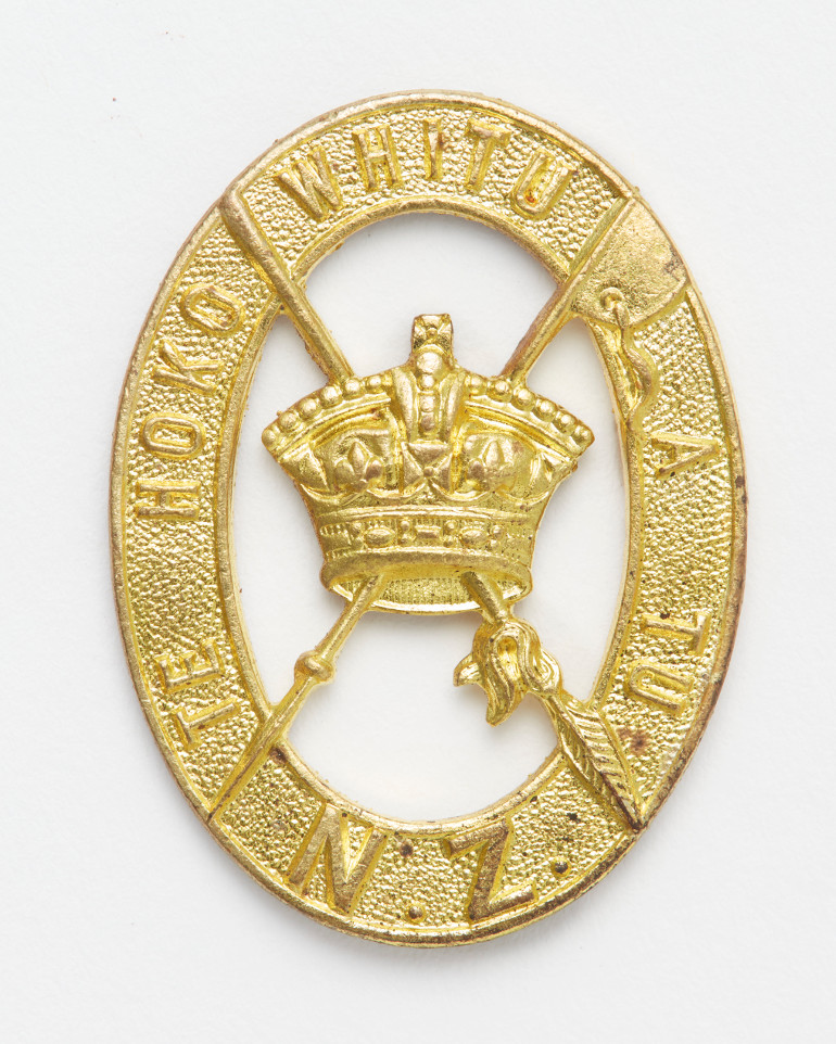 Brass oval cap badge with a taiaha and tewhatewha crossing through a crown at the centre and the words 'TE HOKO WHITU A TU. NZ' around the outside.