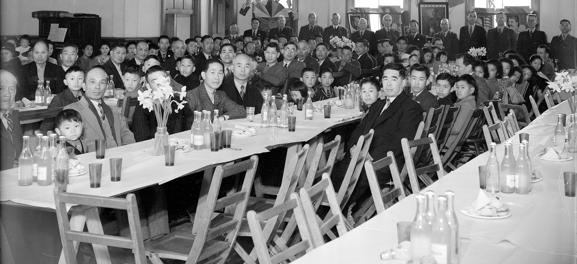 Indoor banquet with long tables set with bottles, glasses, and flower centrepieces, rows of chairs and a large, seated audience.