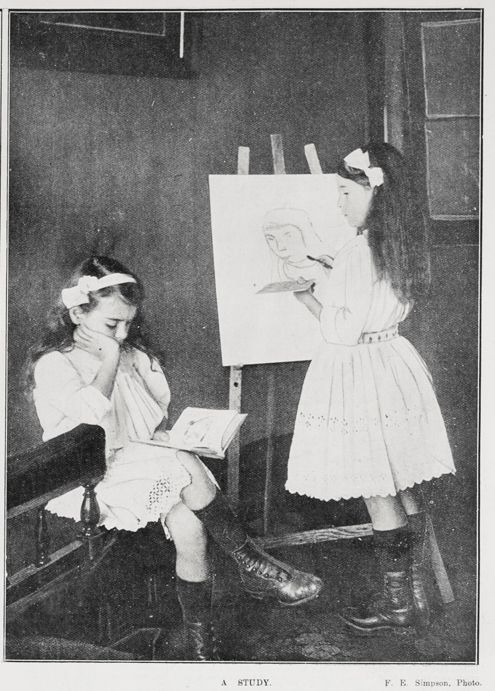 A young girl painting another girl who is seated wearing a book. They wear white dresses and bows, and black boots. 