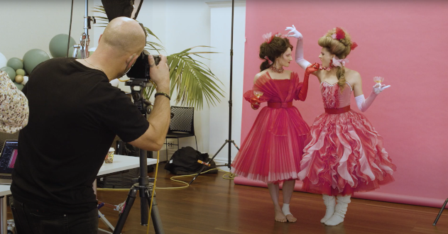 Two people in vintage pink dresses pose for a photographer with drinks in a studio set with a pink backdrop, green balloons, and plants.