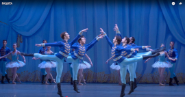 Ballet dancers in blue military-style costumes perform a synchronised pose on stage, with dancers in white tutus in the background.