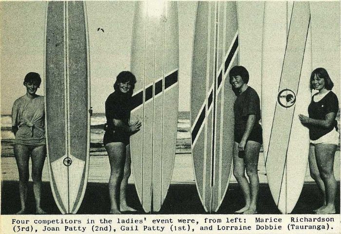 Four young women standing next to 1960s era surfboards.