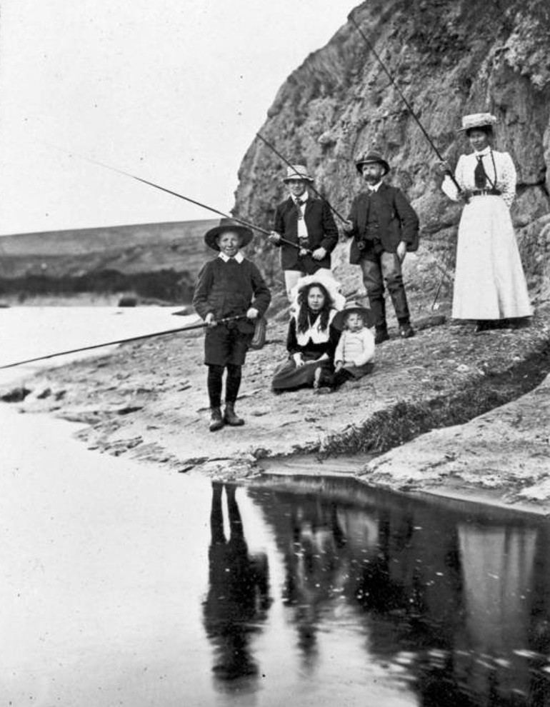 Group of people wearing Edwardian style clothing, standing near waterand holding fishing rods.