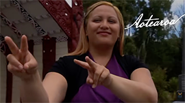 Person signing in New Zealand Sign Language in front of a Māori meeting house, with the word 'Aotearoa' written in white script.