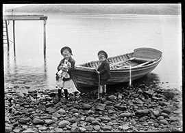 Two young children stand on a rocky beach beside a small wooden boat pulled up on the shore, with calm water behind them and a simple jetty extending into the distance.
