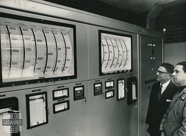 Two individuals standing in front of a large control panel with multiple gauges, meters, and two large curved scale displays.