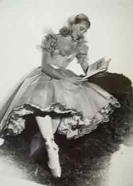 A woman wearing a ballet costume with a layered skirt and pointe shoes, seated and reading a book.