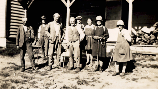 A group of people wearing 1920s-era clothing and walking sticks standing in front of a hut.