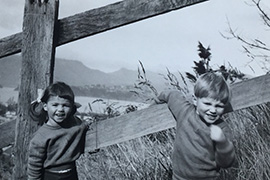 Two young smiling children leaning on a wooden fence with long grass and a harbour behind them.
