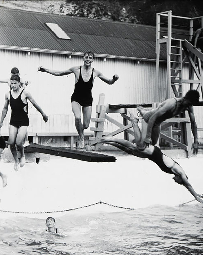 A group of men in black one-piece swimsuits jumping off a diving board into a pool.