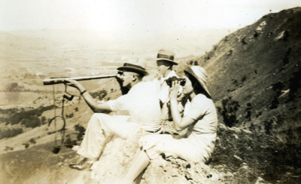 Two men and a woman sitting on a hill. One of the men is looking through a handheld telescope, and the woman is using a pair of binoculars.