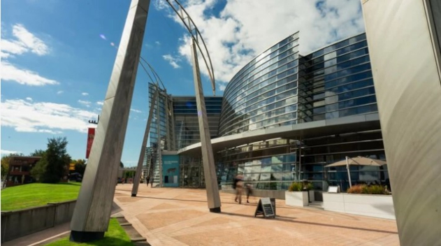 Modern multi-storey building with a curved glass facade, metal supports, and a walkway leading to the entrance.