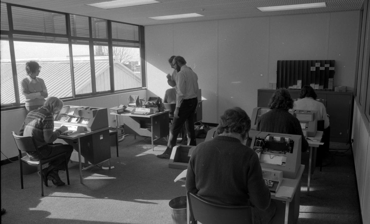 An office room with people talking and working on 1970s era computer machines. 