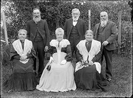 Six people pose outdoors in front of dense foliage, with three seated in front and three standing behind, all dressed in formal early 20th-century clothing with high-collared garments and jackets.