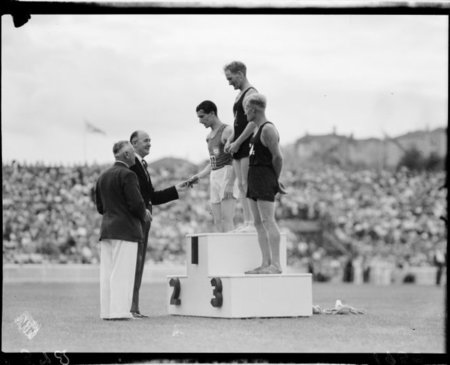 Three men wearing singlets and shorts standing on an outdoors podium, one receiving an award from another man wearing a suit.