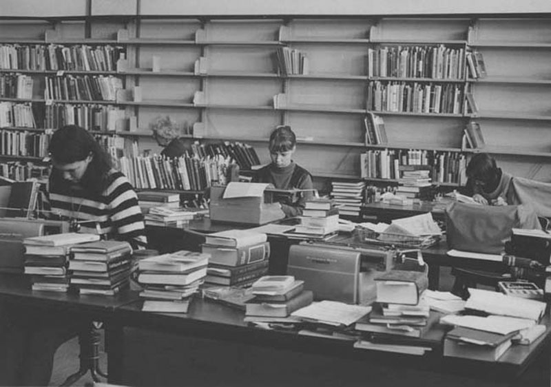 Three people working at large desks surrounded by piles of books and half empty book shelves.
