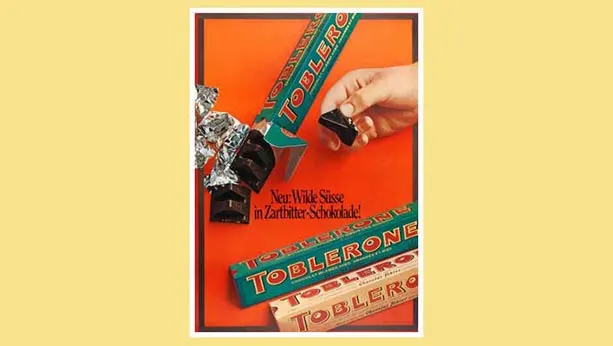 Ad from 1969 of Dark chocolate Toblerone. 2 packets of dark chocolate Toblerone - 1 open with a hand holding a piece from that package. 1 original flavour package unopened.