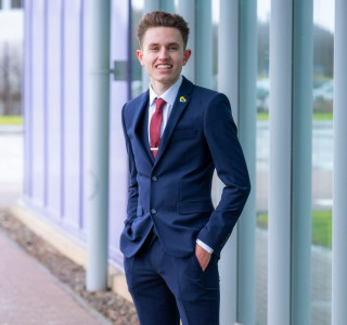 Young man in a navy suit stands with his hands in his pockets and smiles to the camera