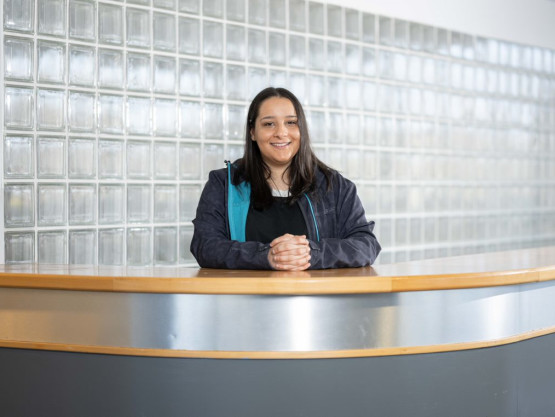 Young woman in a blue jacket stands behind a reception desk with a glass wall in the background