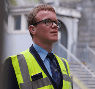 A young man in glasses, wearing a high-vis jacket stands in front of stairs