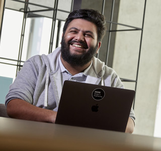 A young man smiles while sitting at a desk with a laptop in front of him.