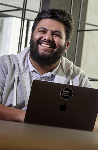 A young man smiles while sitting at a desk with a laptop in front of him.