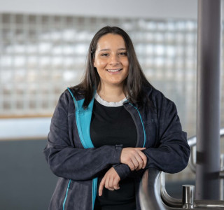 Young woman in a blue jacket leans on a railing while smiling into the camera