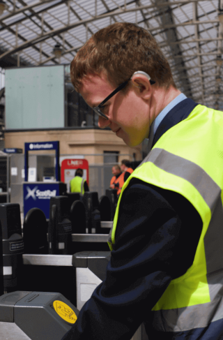 A young man in a high-vis jacket operates a ticket barrier in a train station