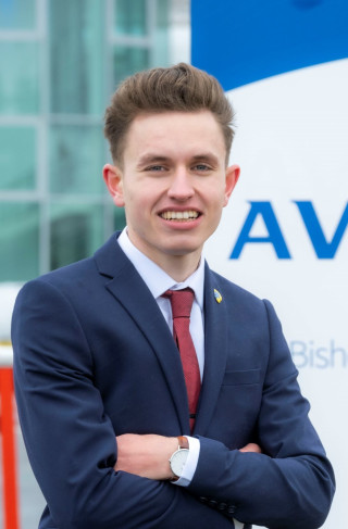 A young man in a navy suit stands in front of the Aviva sign with his arms crossed and smiles to the camera