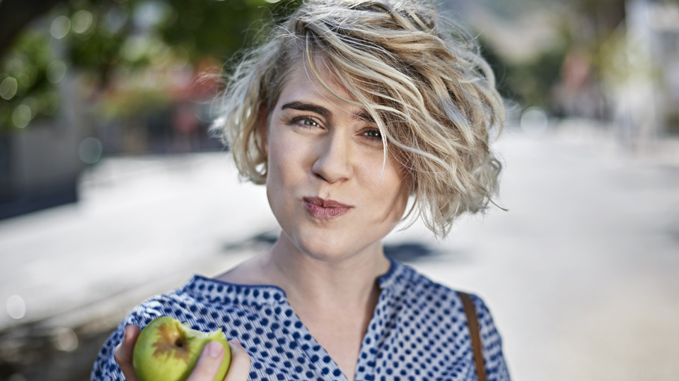 A young woman smiles at the camera as she eats a green apple.