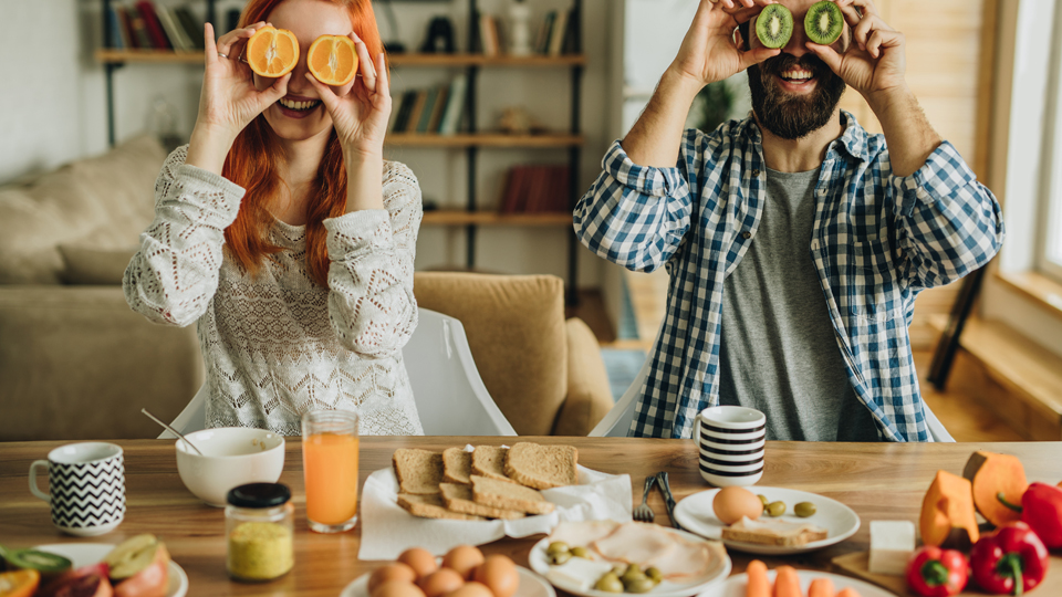 A couple laughing as they hold slices of fruit in place of their eyes at the breakfast table