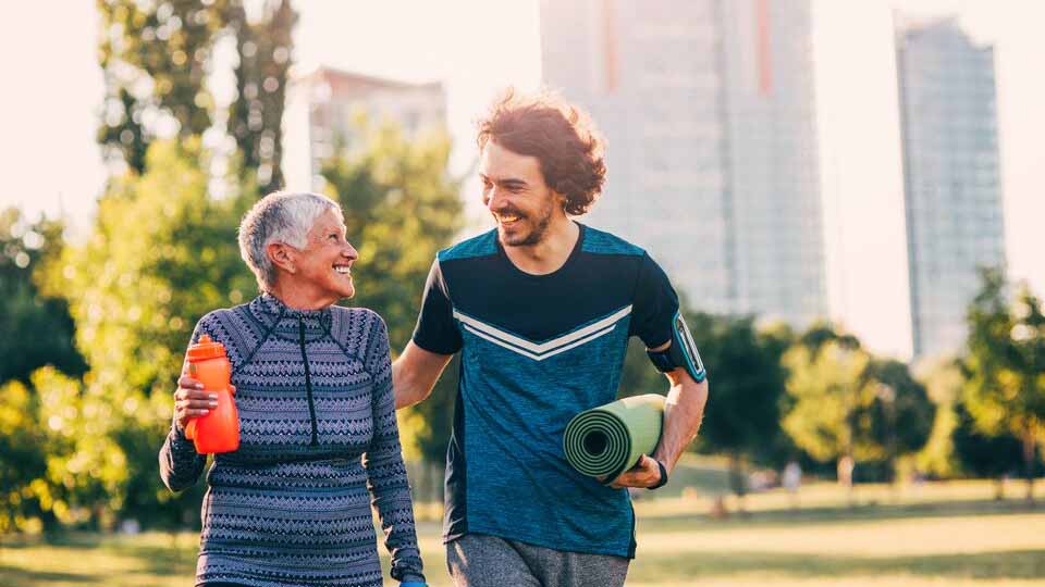 A smiling young man walks and pats a laughing senior woman on the back while holding a yoga mat.