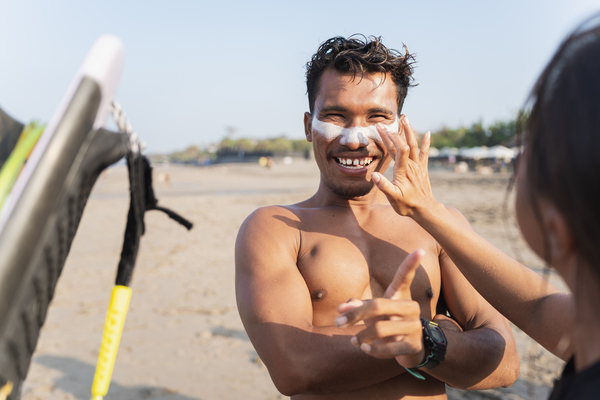 Woman on a beach applying sunscreen to the face of a man with a surfboard next to him.